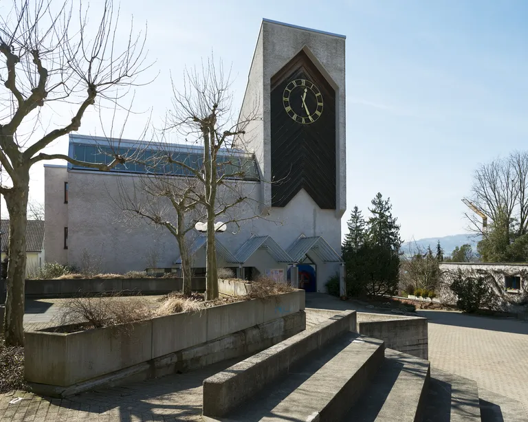 Kirche Oberengstringen mit modernem Turm und grosser Uhr, umgeben von kahlen Bäumen und Treppen an einem sonnigen Tag.