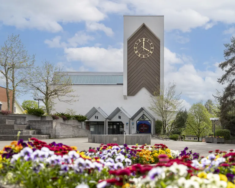 Kirche Oberengstringen mit modernem Turm und grosser Uhr, umgeben von kahlen Bäumen und Treppen an einem sonnigen Tag.