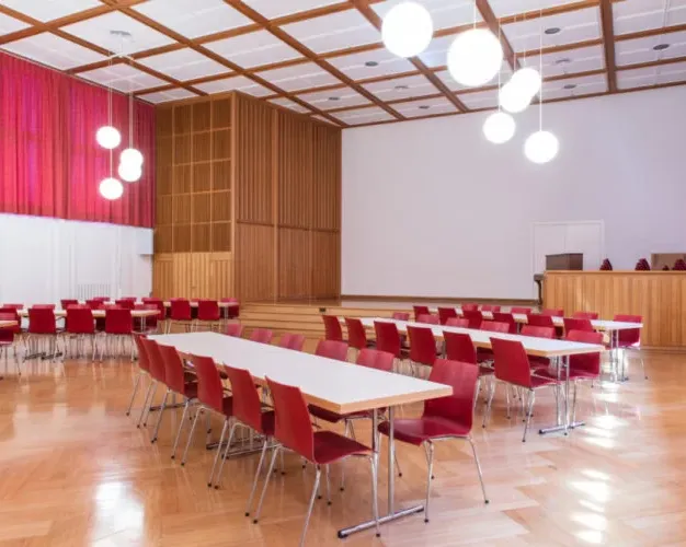 Heller Saal im Kirchgemeindehaus Oberengstringen mit Holzboden, roten Stühlen und langen Tischen, moderne Lampen hängen von der Decke.