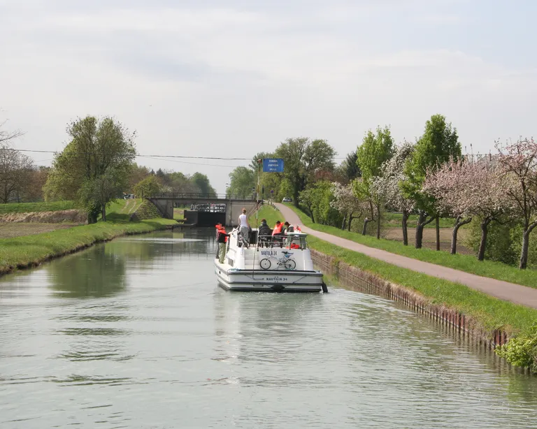 Ein Boot mit Menschen fährt auf einem ruhigen Kanal, umgeben von Bäumen und einem Weg am Ufer.