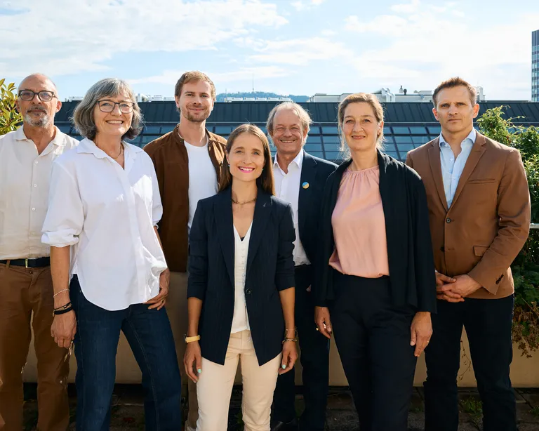 Sieben Menschen stehen im Freien auf einer Terrasse, im Hintergrund sind Gebäude sichtbar.