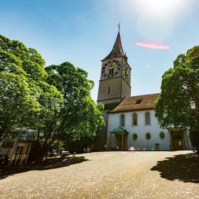 Historische Kirche mit Turm, umgeben von Bäumen auf einem sonnigen Platz.