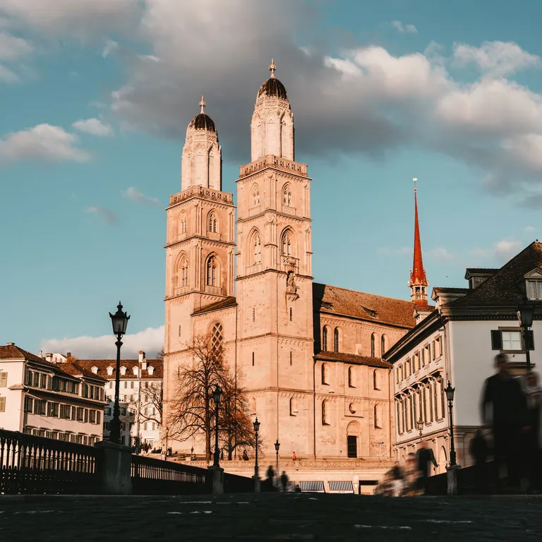 Das Grossmünster mit seinen zwei Türmen bei Sonnenuntergang, umgeben von historischen Gebäuden und blauem Himmel mit Wolken.