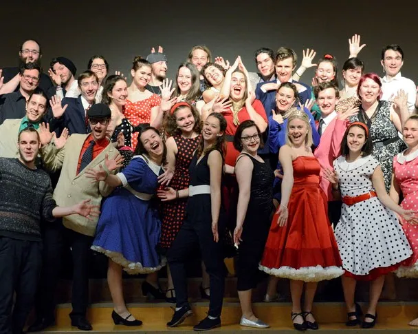 Eine grosse, fröhliche Gruppe vom Musicalprojekt in Zürich posiert gemeinsam auf einer Bühne, viele tragen farbige, festliche Kleider.