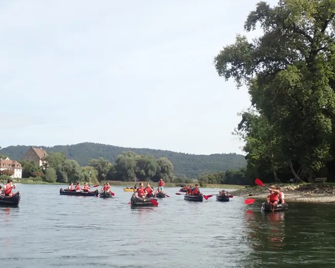Mehrere Teilnehmende des Kurses für Jugendleiterinnen in Kanus auf einem ruhigen Fluss, im Hintergrund Bäume und Häuser, rechts ein grünes Ufer.