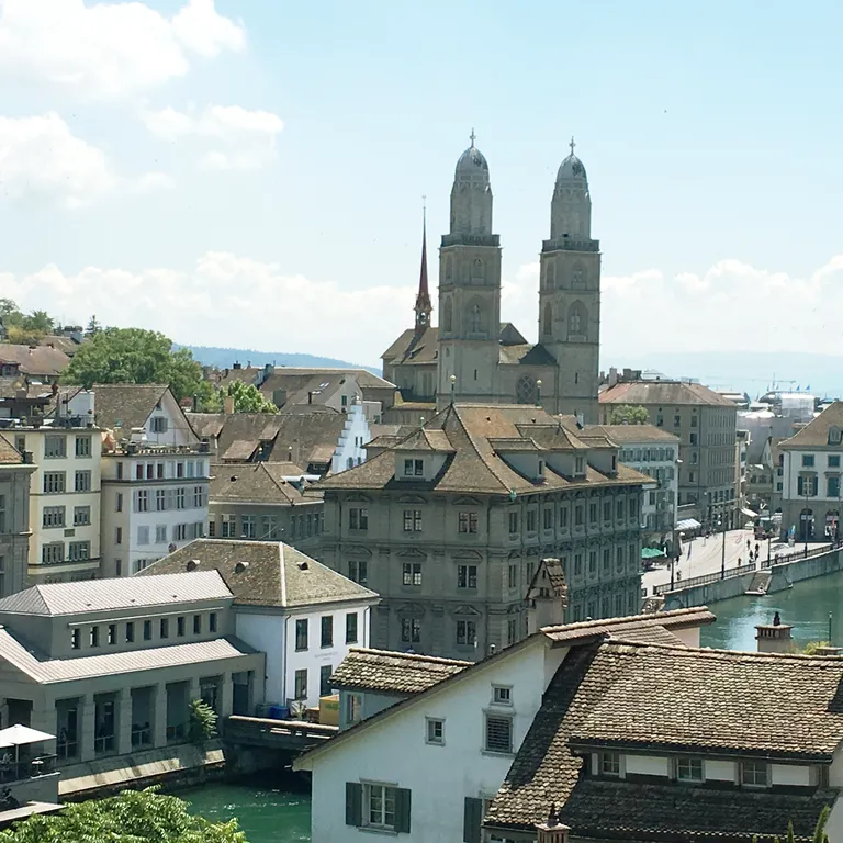 Blick über Zürich mit dem Grossmünster mit seinen zwei Türmen im Hintergrund, dem Rathaus im Vordergrund und der Limmat unter blauem Himmel.