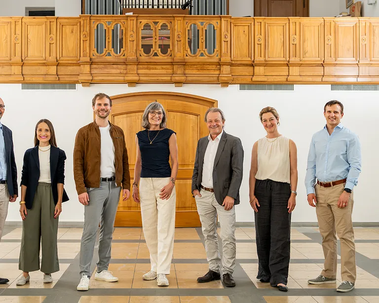 Sieben Menschen stehen nebeneinander in einem hellen Raum mit Holzgalerie und Orgel im Hintergrund.