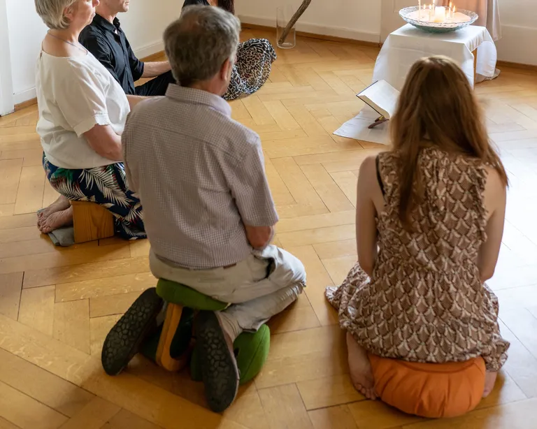 Fünf Personen sitzen meditierend auf Kissen in einem hellen Raum, vor einem kleinen Altar mit Tüchern und Ästen.