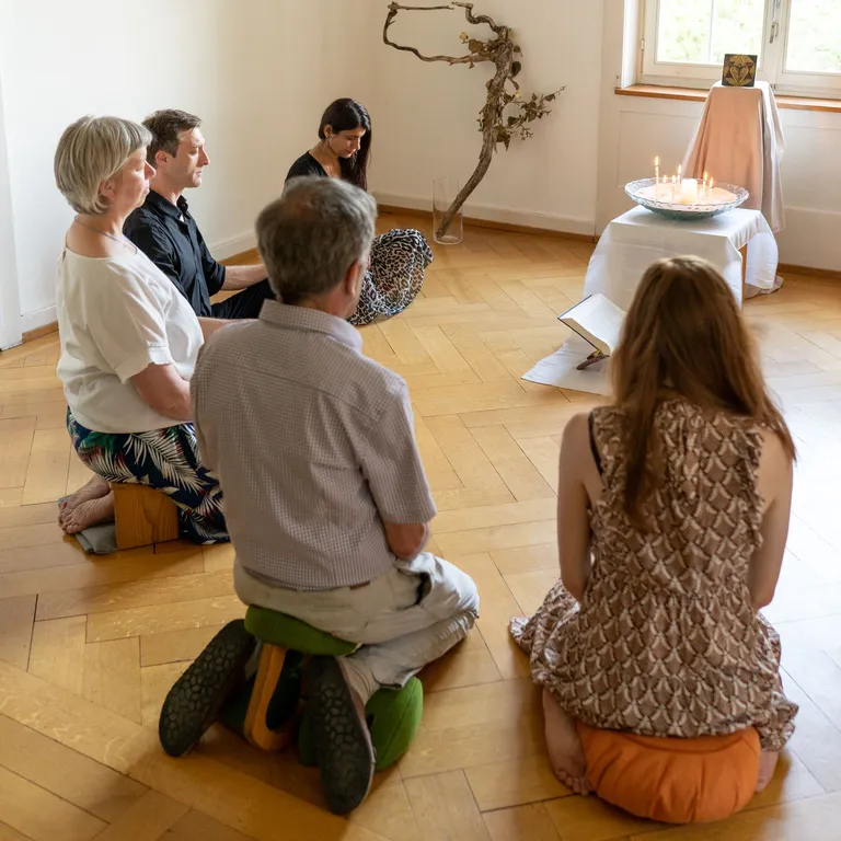 Fünf Personen sitzen meditierend auf Kissen in einem hellen Raum, vor einem kleinen Altar mit Tüchern und Ästen.