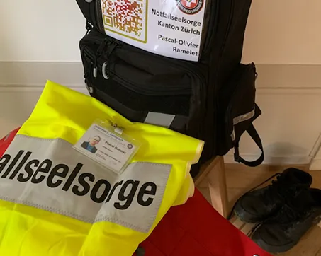 Ein gelbe Weste mit der Aufschrift «Notfallseelsorge» liegt auf einem Rucksack, daneben stehen schwarze Schuhe auf dem Boden.