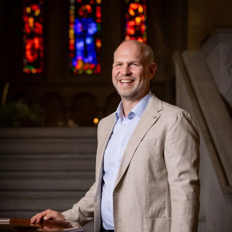 Pfarrer Christian Walti  steht im hellen Sakko in einer Kirche vor einer Treppe, im Hintergrund sind bunte Kirchenfenster zu sehen.