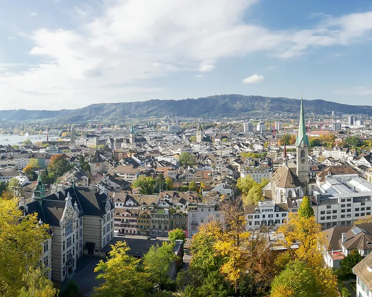 Panorama der Stadt Zürich mit vielen Häusern, einer Kirche mit hohem Turm und umliegenden Hügeln unter blauem Himmel.